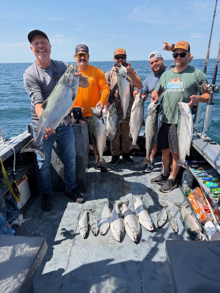 Cinq pêcheurs souriants à bord du bateau avec leurs saumons, d’autres prises alignées sur le pont