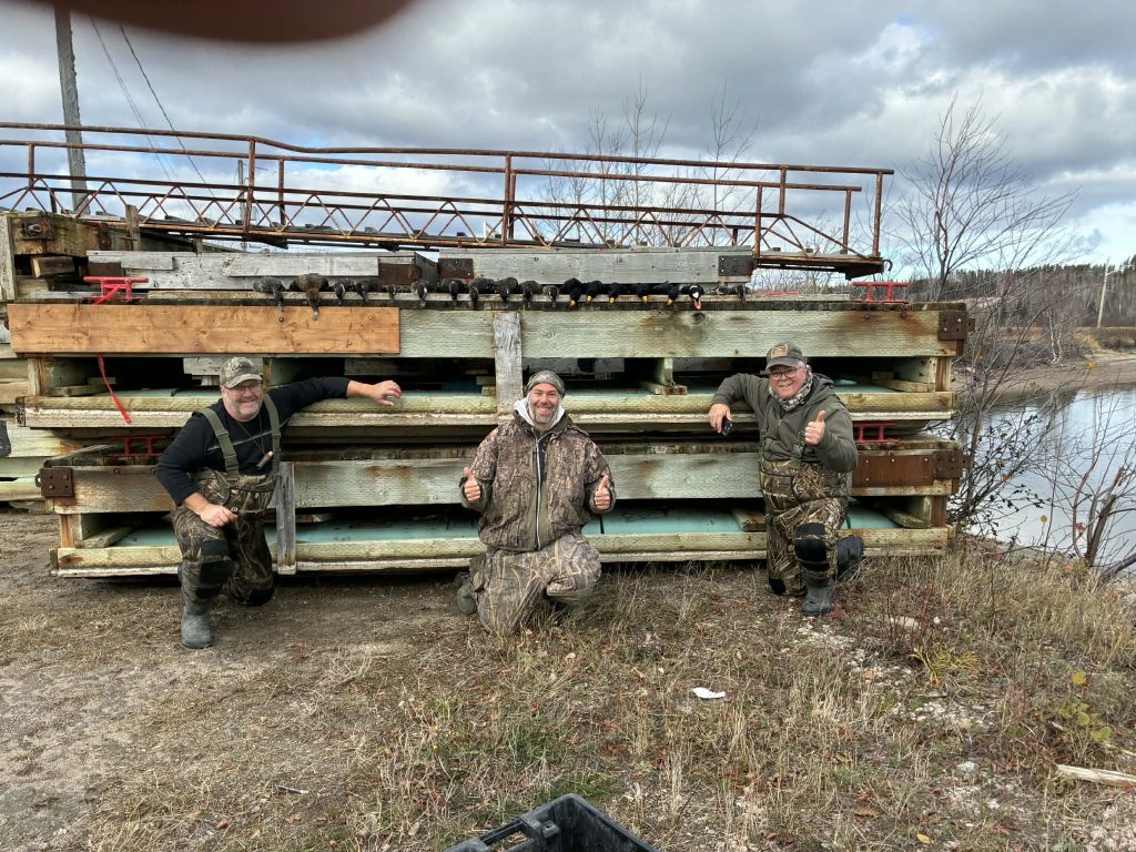 Trois chasseurs en camouflage devant les structures de caches empilées au bord de l’eau