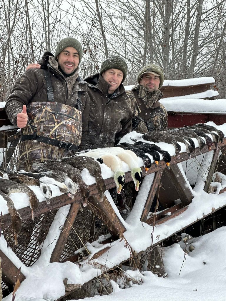 Trois chasseurs souriants derrière une rangée de garrots et de harles posés sur une structure de bois enneigée
