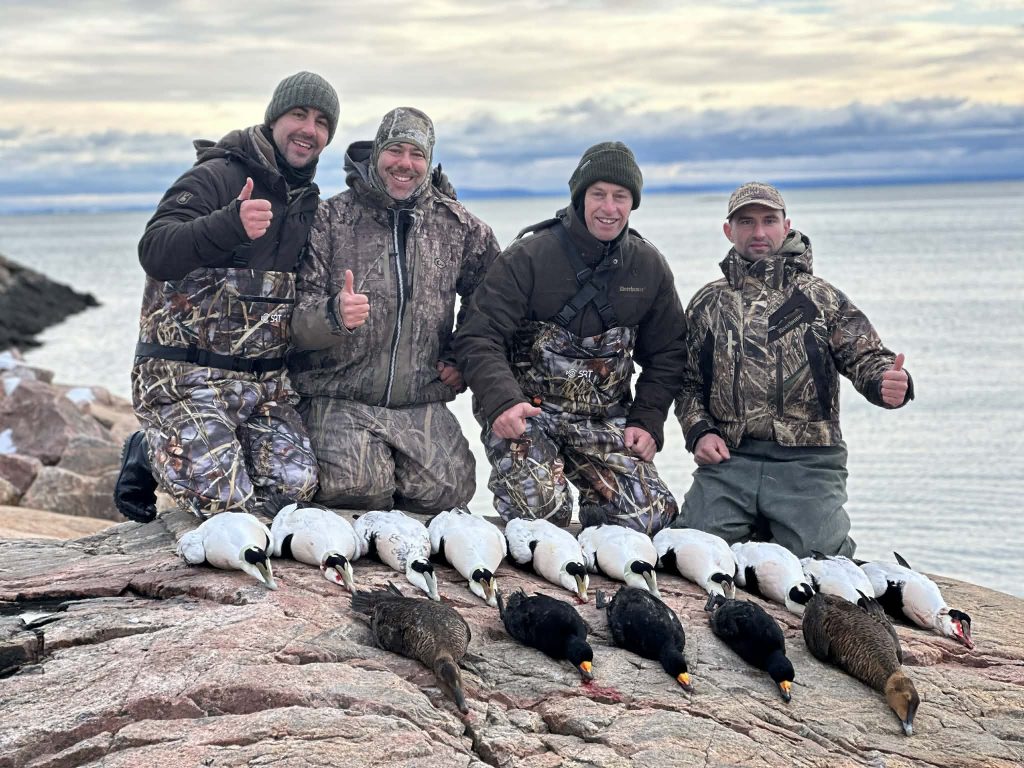Quatre chasseurs souriants sur les rochers derrière une rangée d’eiders et de macreuses