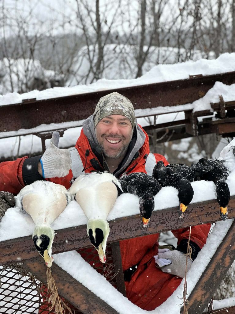 Chasseur souriant sous la neige derrière deux eiders et deux macreuses posés sur une rampe